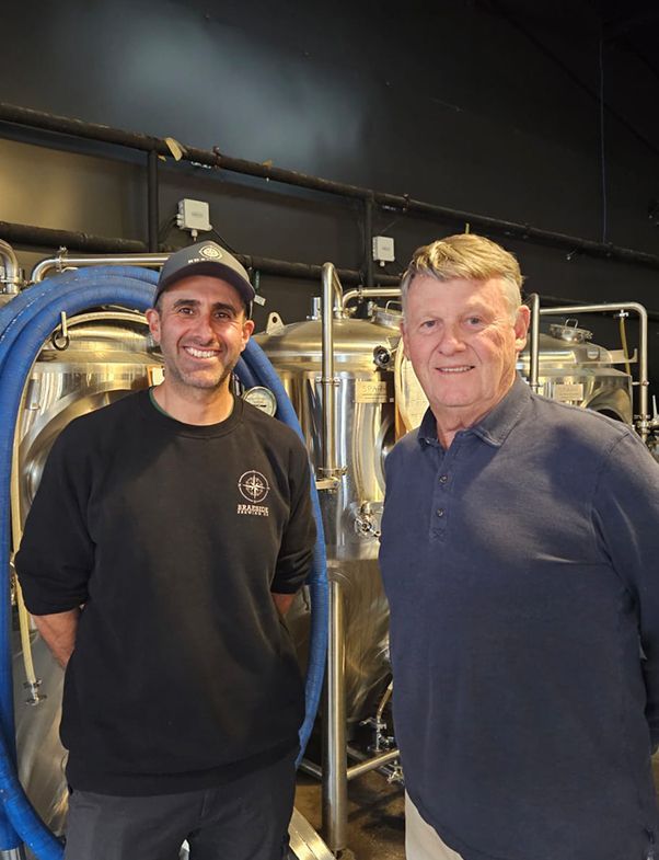 Two men stand in a brewery, in front of brewing equipment. One in a baseball cap and black shirt smiles. The other wears a blue shirt.
