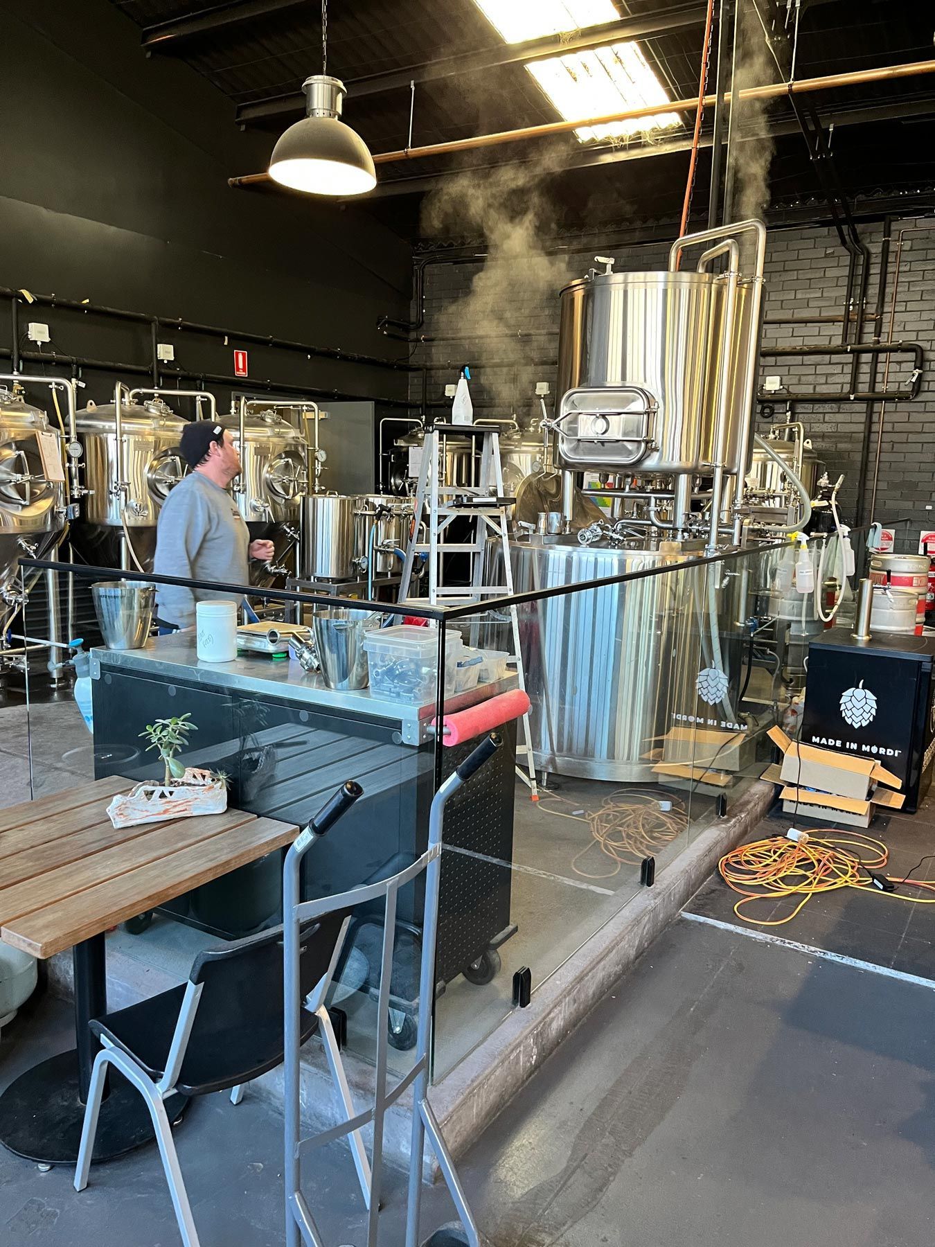 Brewery interior: man near stainless steel tanks with steam. Wooden table and chairs in foreground.