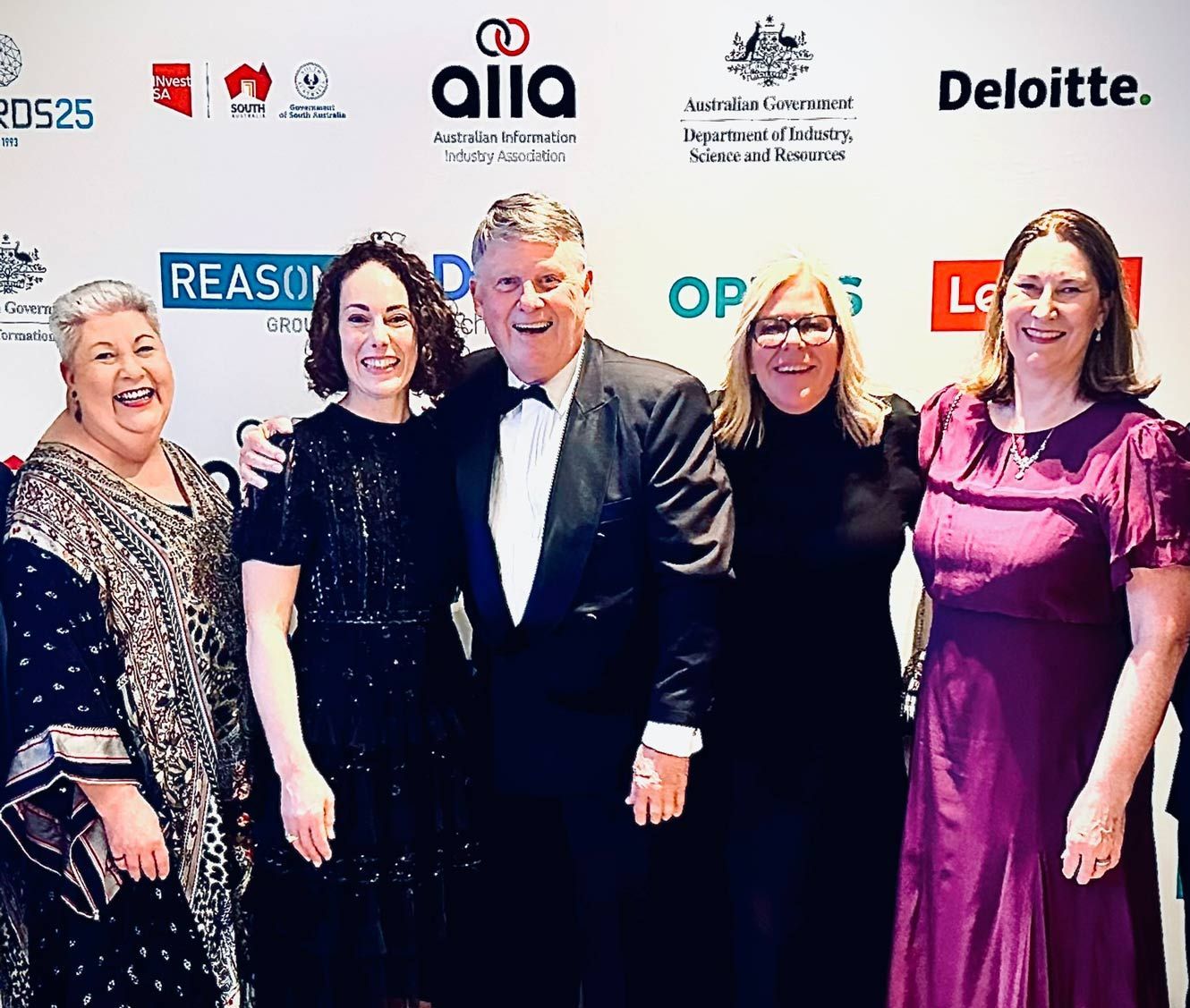 Five people posing for a photo. A man in a tuxedo is in the center with arms around two women. Other women on either side smile.