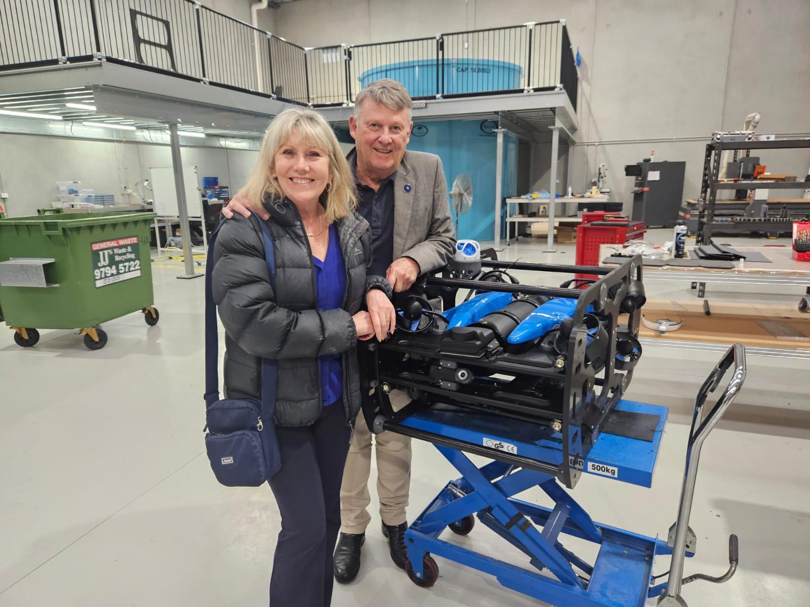 Woman and man pose next to a black machine on a lift, indoors. Woman in black jacket, man in blazer.
