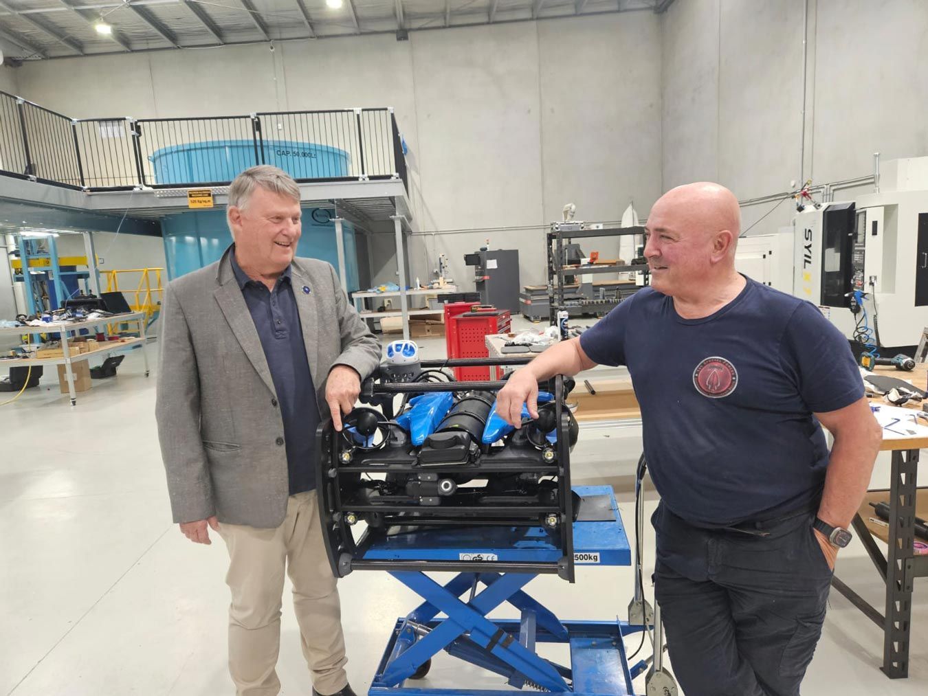 Two men beside a blue engine on a lift table in a workshop, one points to it.