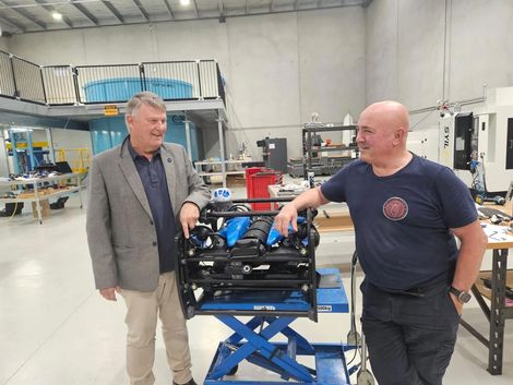 Two men beside a blue engine on a lift table in a workshop, one points to it.