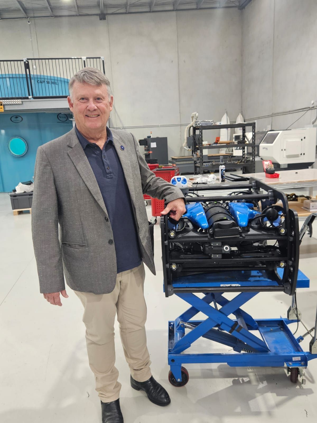 Man in a blazer standing next to a large blue engine on a blue lift table in a workshop.