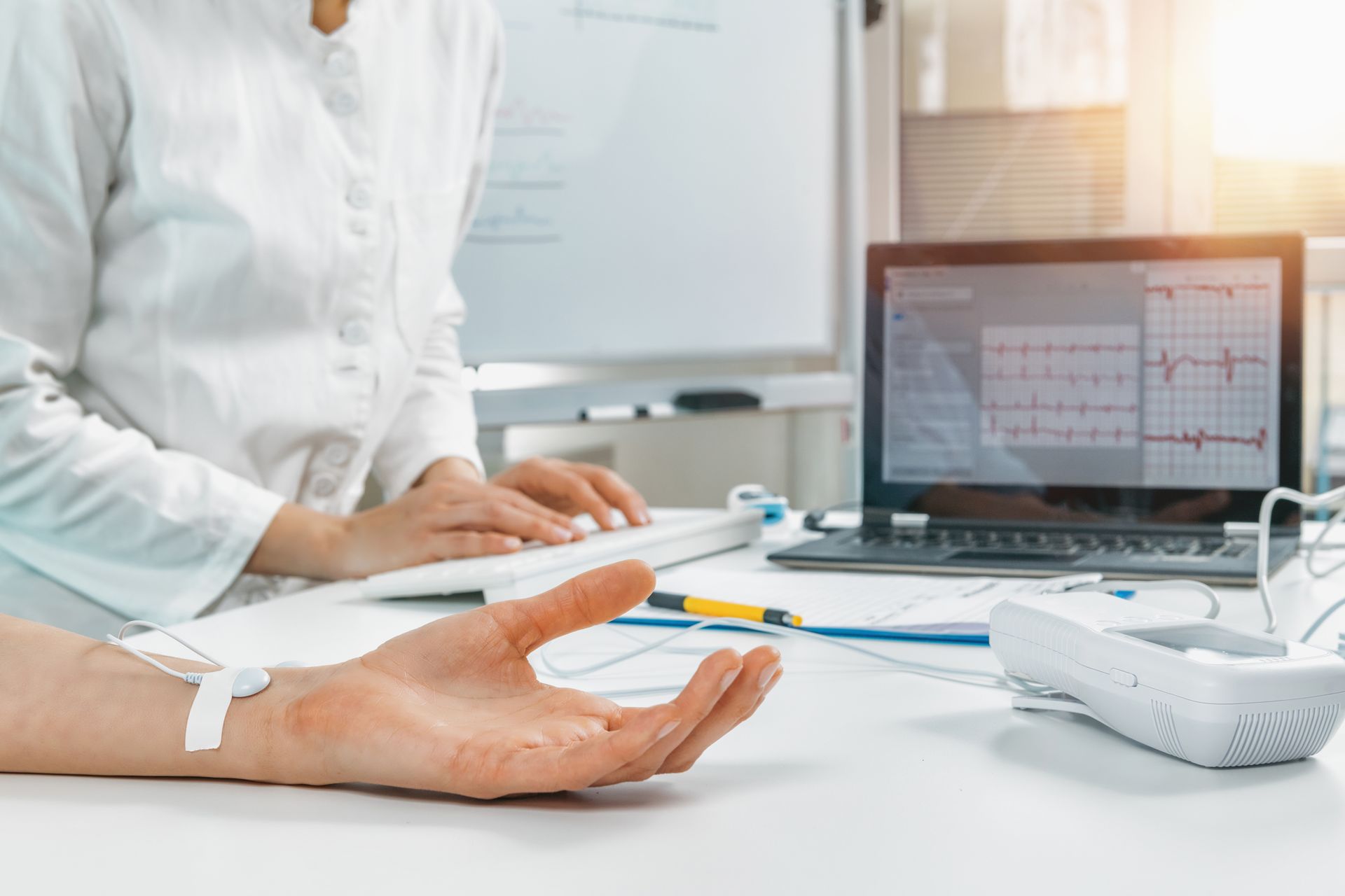 Der Arm einer Person ist mit einem medizinischen Gerät verbunden, während eine andere Person an einem Computer arbeitet, der ein Herzrhythmusdiagramm anzeigt.