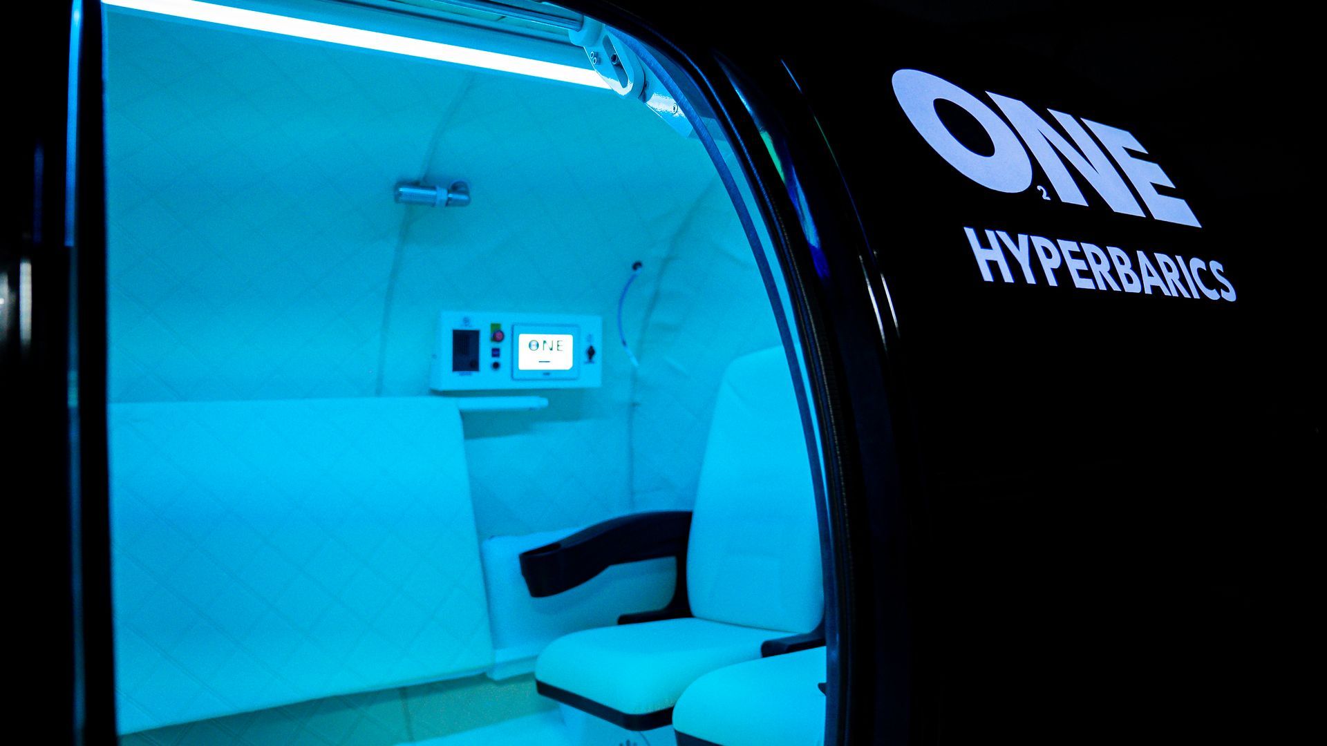Innenansicht einer Überdruckkammer, blau beleuchteter Innenraum, weißer Sitz und Bedienfeld mit dem Logo „ONE HYPERBARICS“.