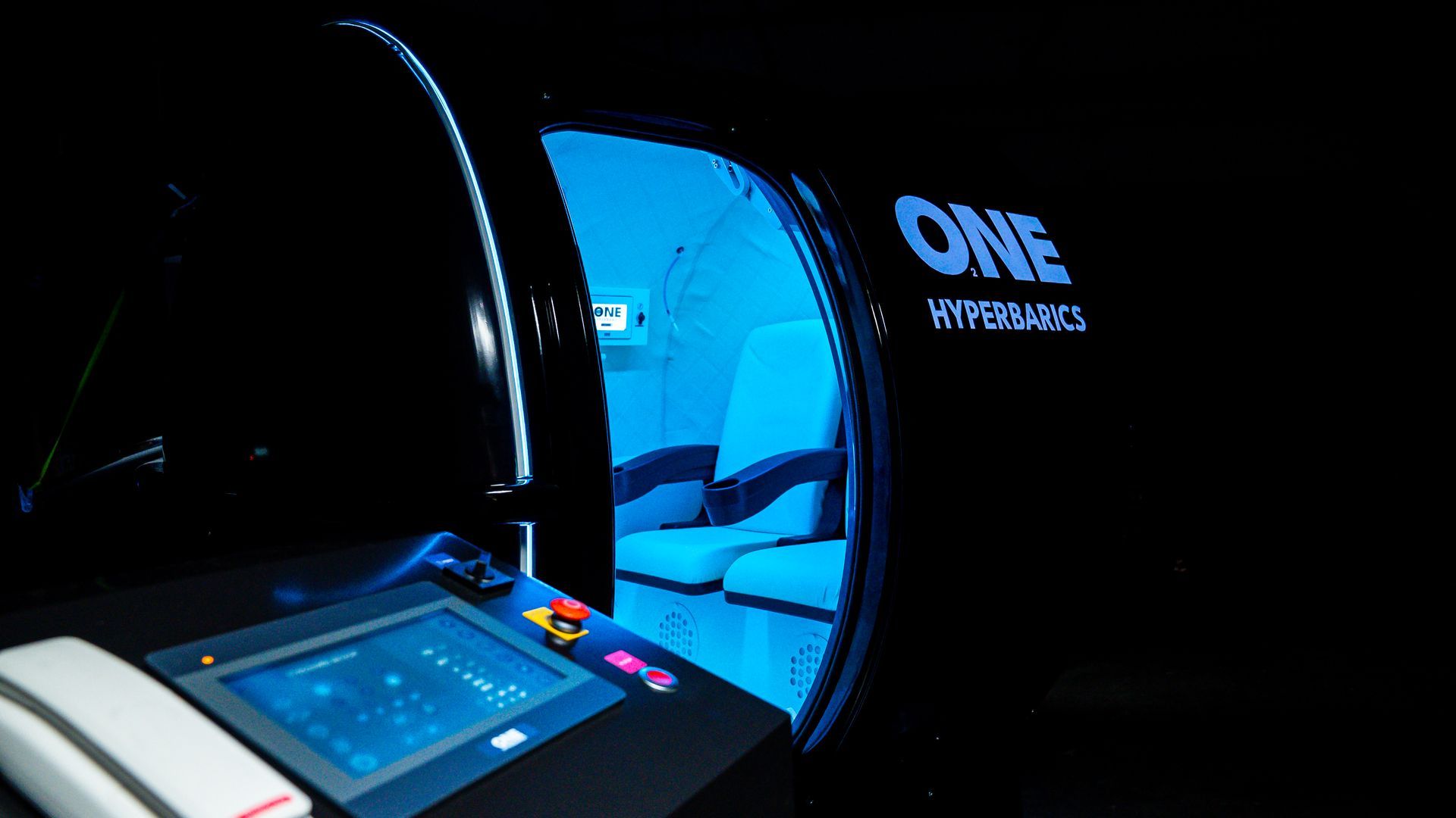 Schwarze Kapsel für HBOT-Behandlung mit blauem Innenraum, Bedienfeld. „ONE HYPERBARICS“-Logo, 