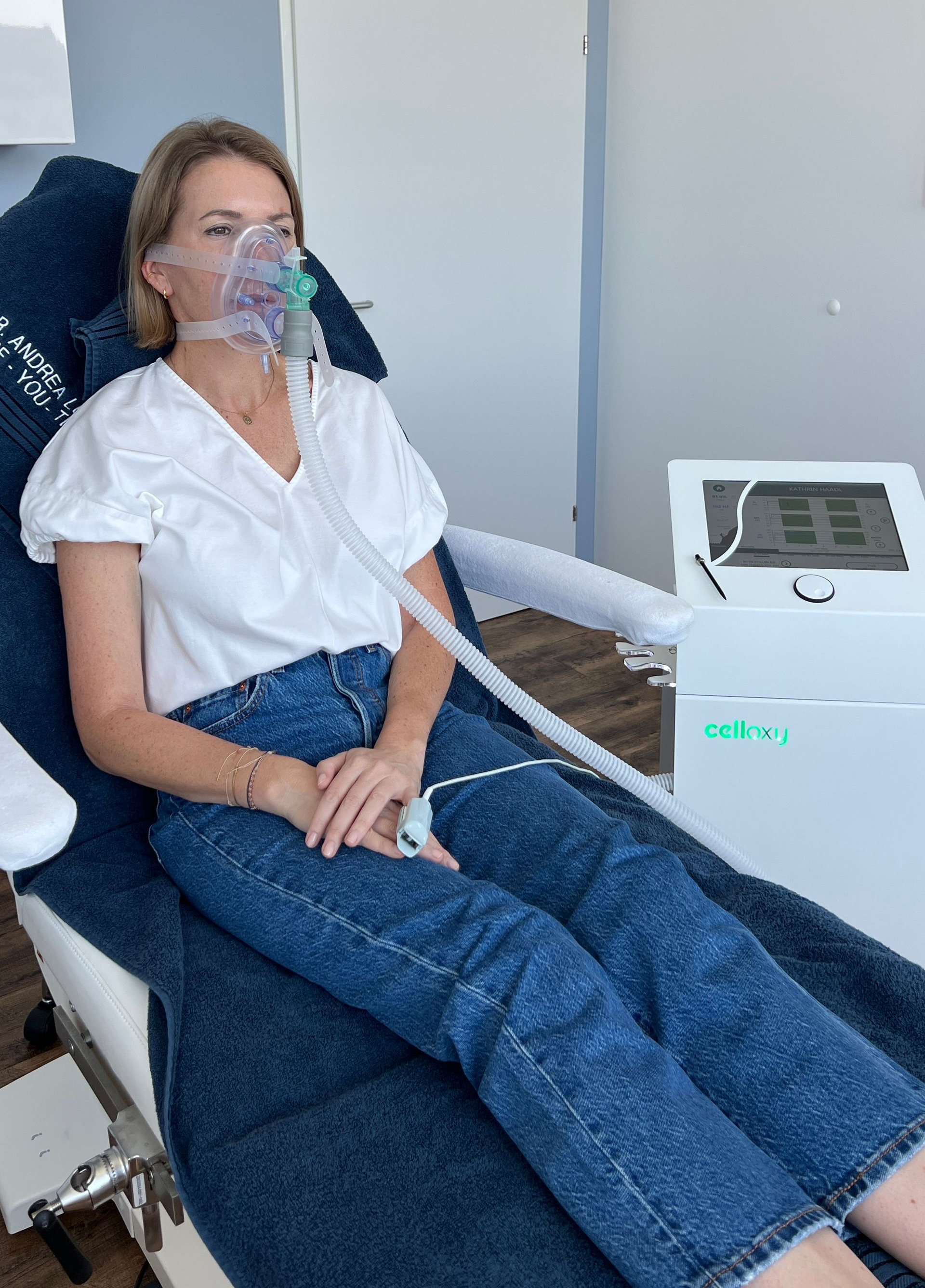 Frau mit Sauerstoffmaske sitzt auf einem Stuhl neben einem Sauerstoffgerät. Drinnen, weißes Hemd, blaue Jeans.