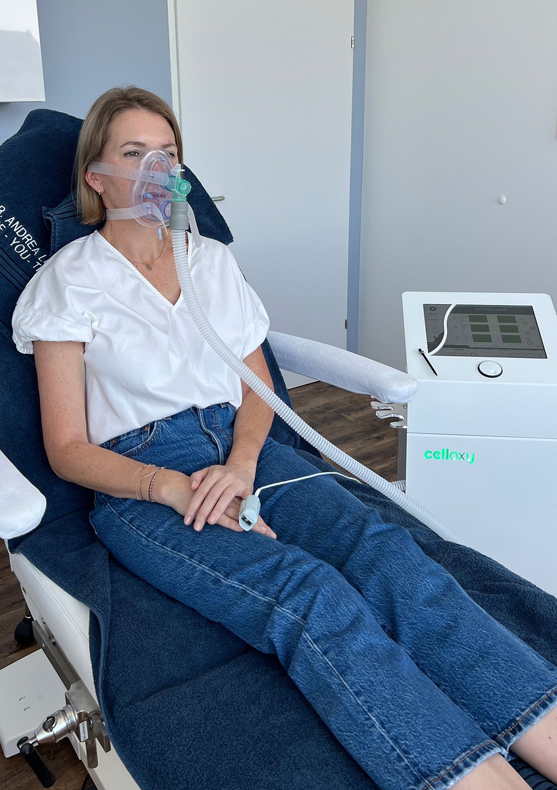 Frau mit Sauerstoffmaske sitzt auf einem Stuhl neben einem Sauerstoffgerät. Drinnen, weißes Hemd, blaue Jeans.