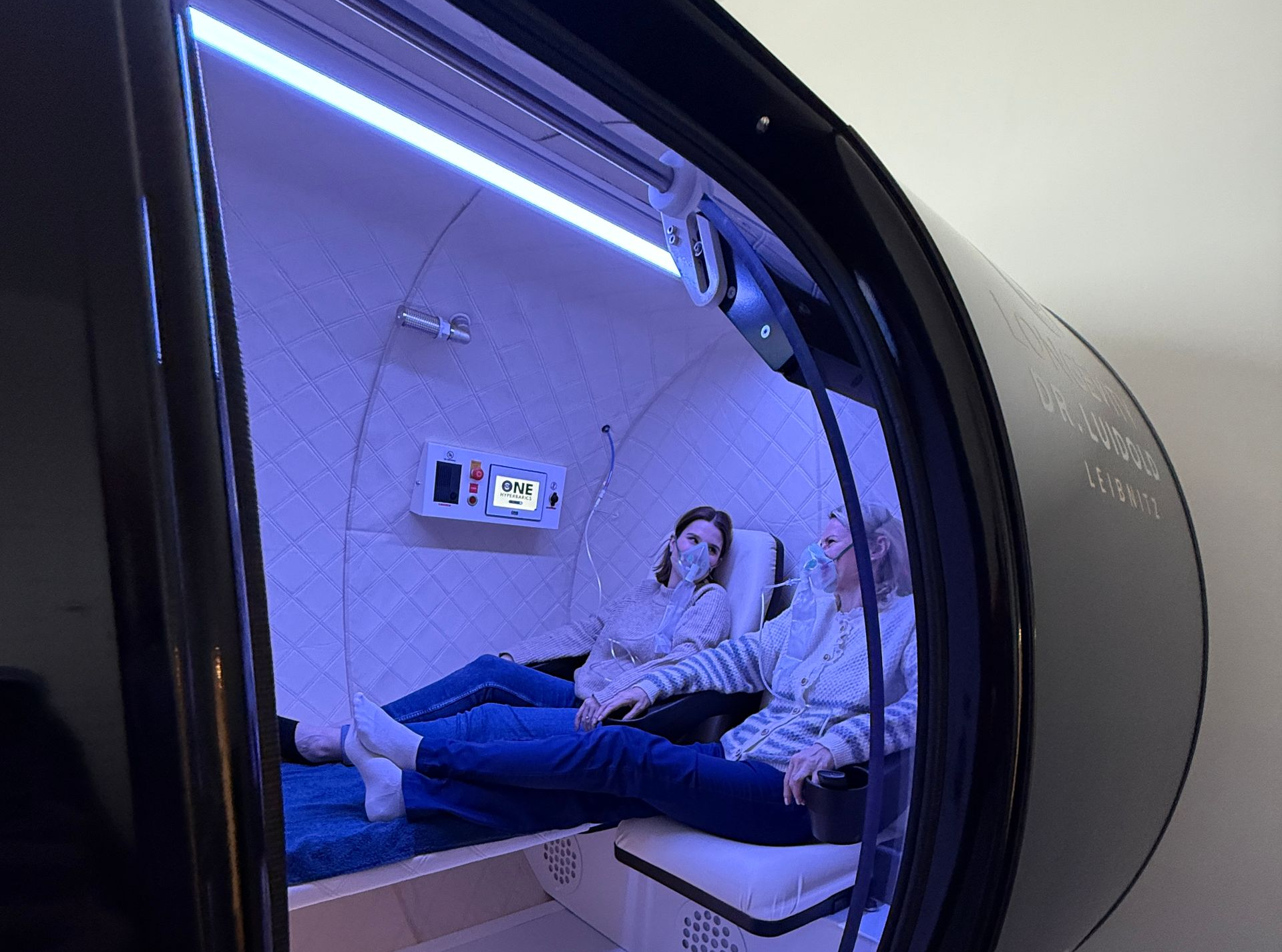 schwarzes Gerät zur HBOT-Behandlung mit der Aufschrift „ONE Fitness Co“ in einem blau beleuchteten Raum.