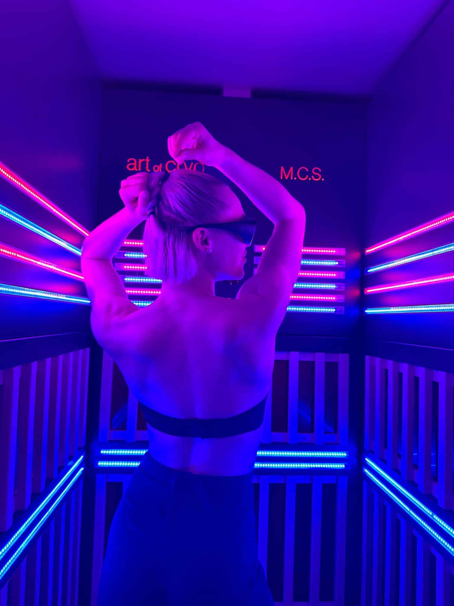 Frau in schwarzer Sportkleidung posiert in neonbeleuchtetem Raum, die Arme erhoben, den Blick von der Kamera abgewandt bei MCS-Therapie