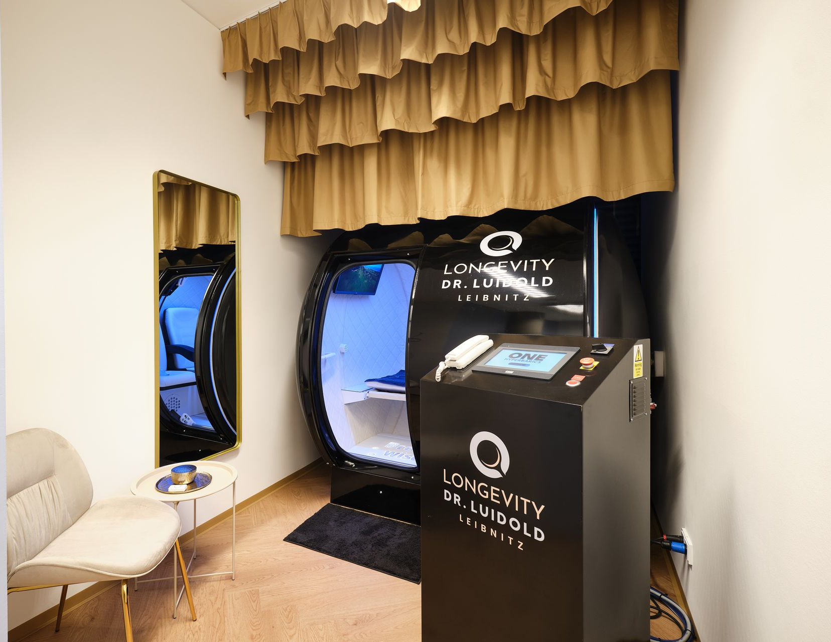 schwarzes Gerät zur HBOT-Behandlung mit der Aufschrift „ONE Fitness Co“ in einem blau beleuchteten Raum.