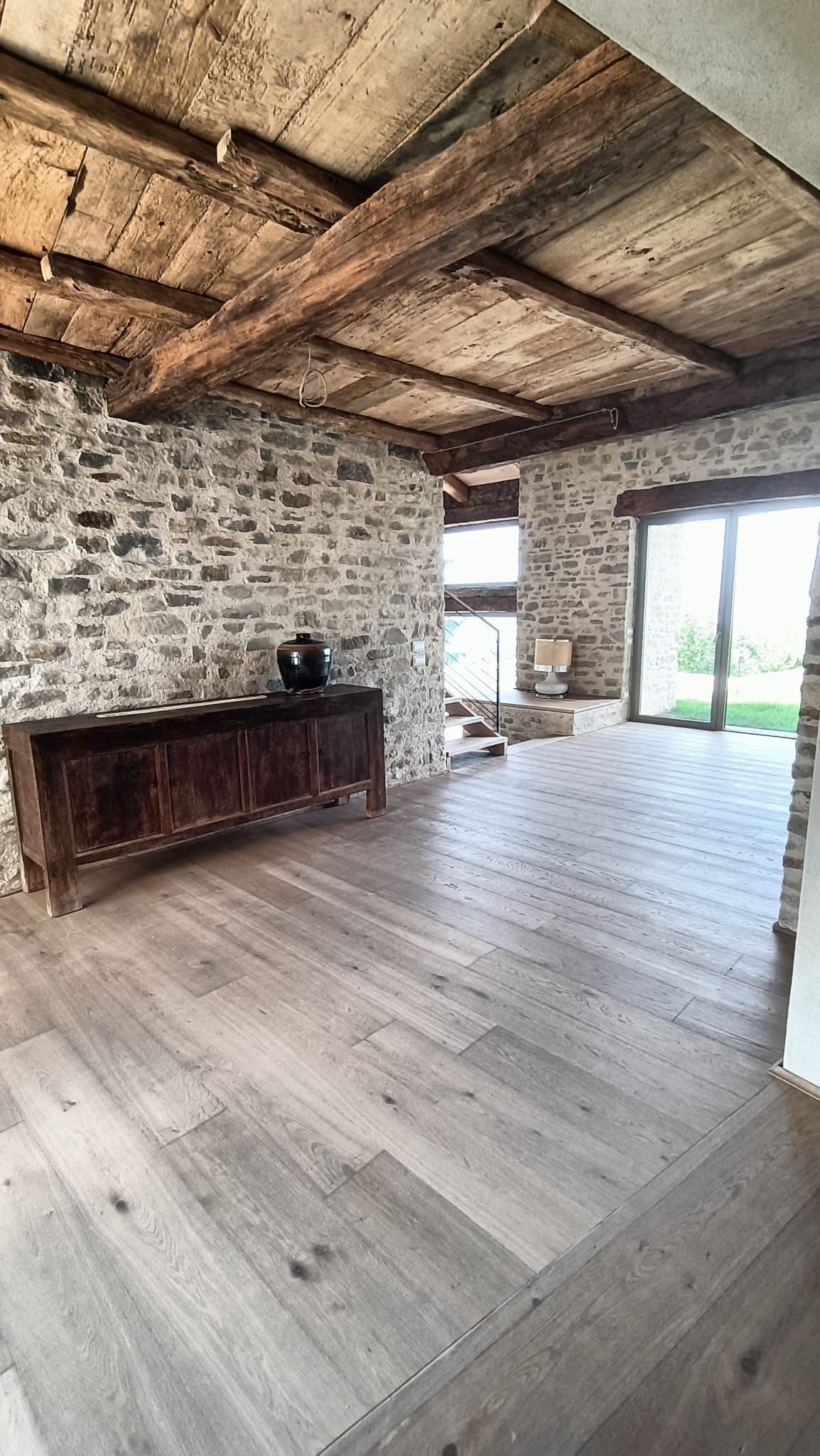 Stanza rustica in pietra con travi in legno a vista, pavimento in assi di legno bianco e un armadio in legno scuro accanto alle grandi finestre.