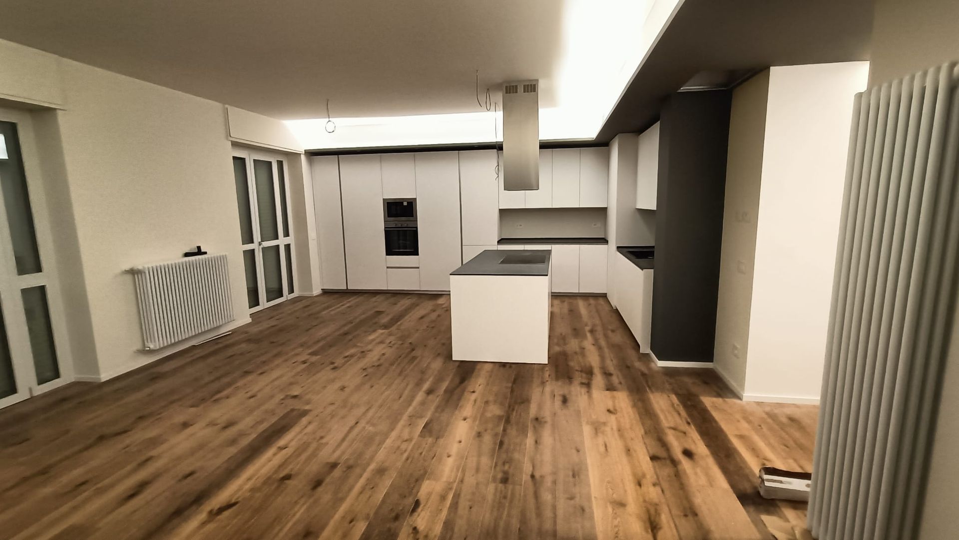 Stanza moderna vuota con pavimento in legno, pareti bianche, termosifoni e un'isola cucina al centro.
