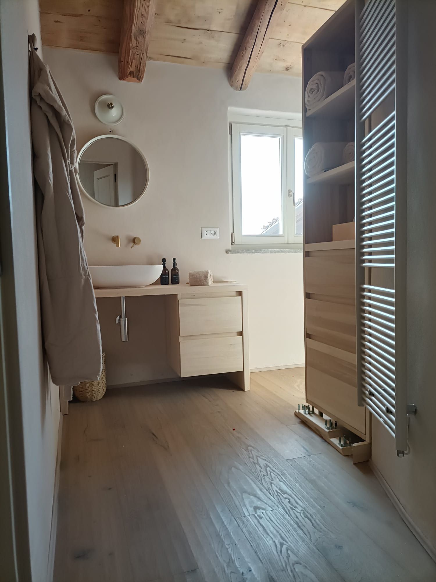 Bagno luminoso con mobile lavabo bianco, specchio rotondo, lucernario, travi in legno e porta dell'armadio a doghe.