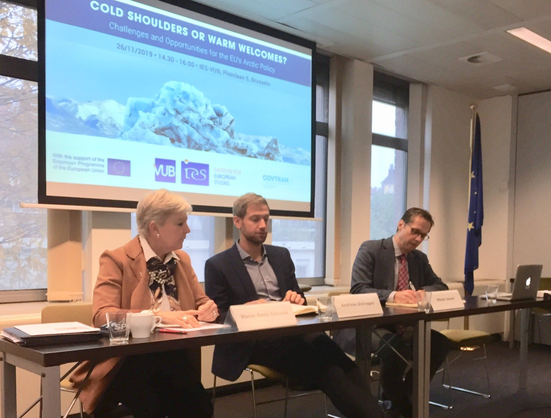 Drei Personen sitzen an einem Tisch vor einem großen Bildschirm.