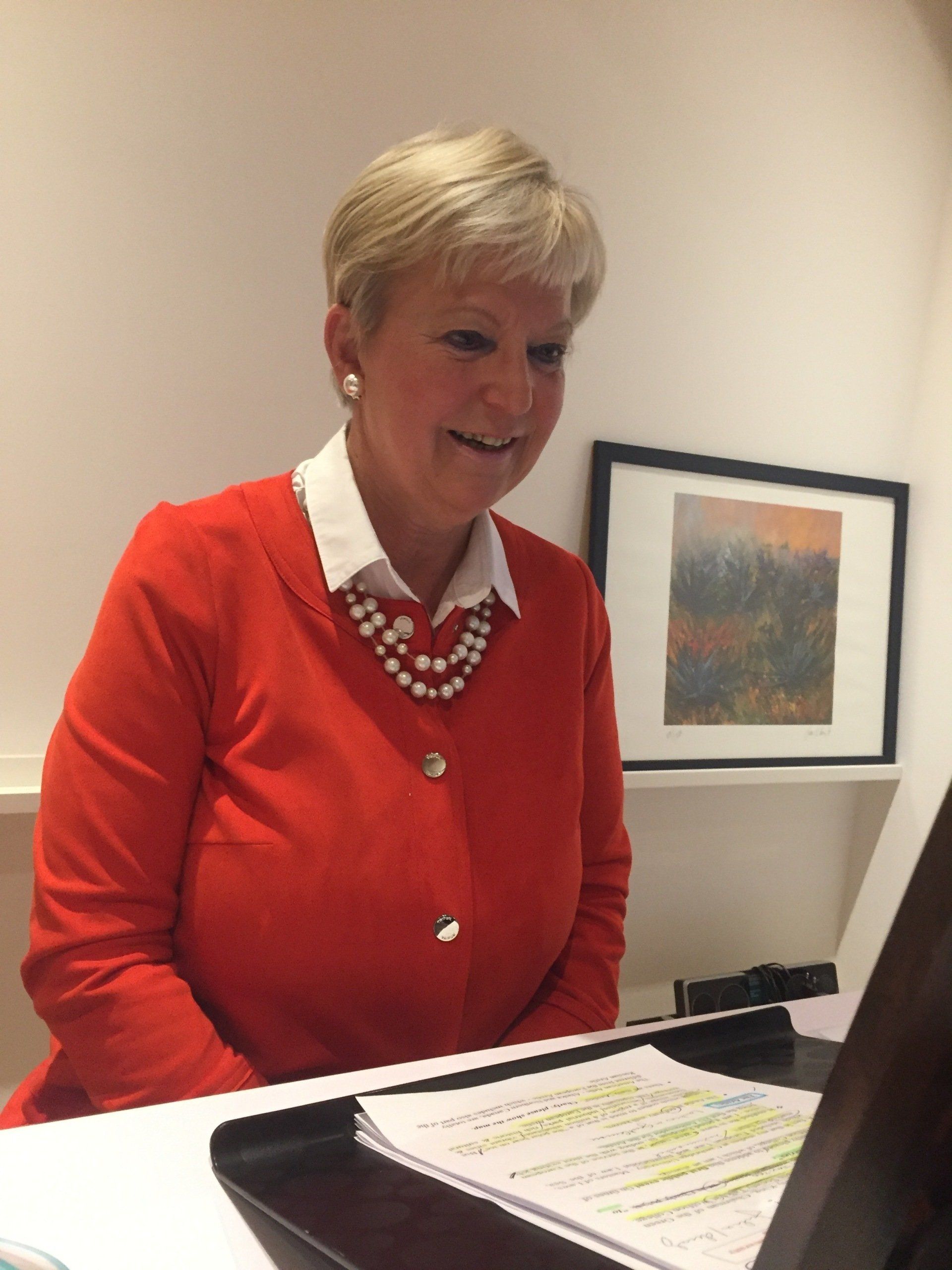 Eine Frau in einem orangefarbenen Pullover sitzt an einem Schreibtisch vor einem Laptop