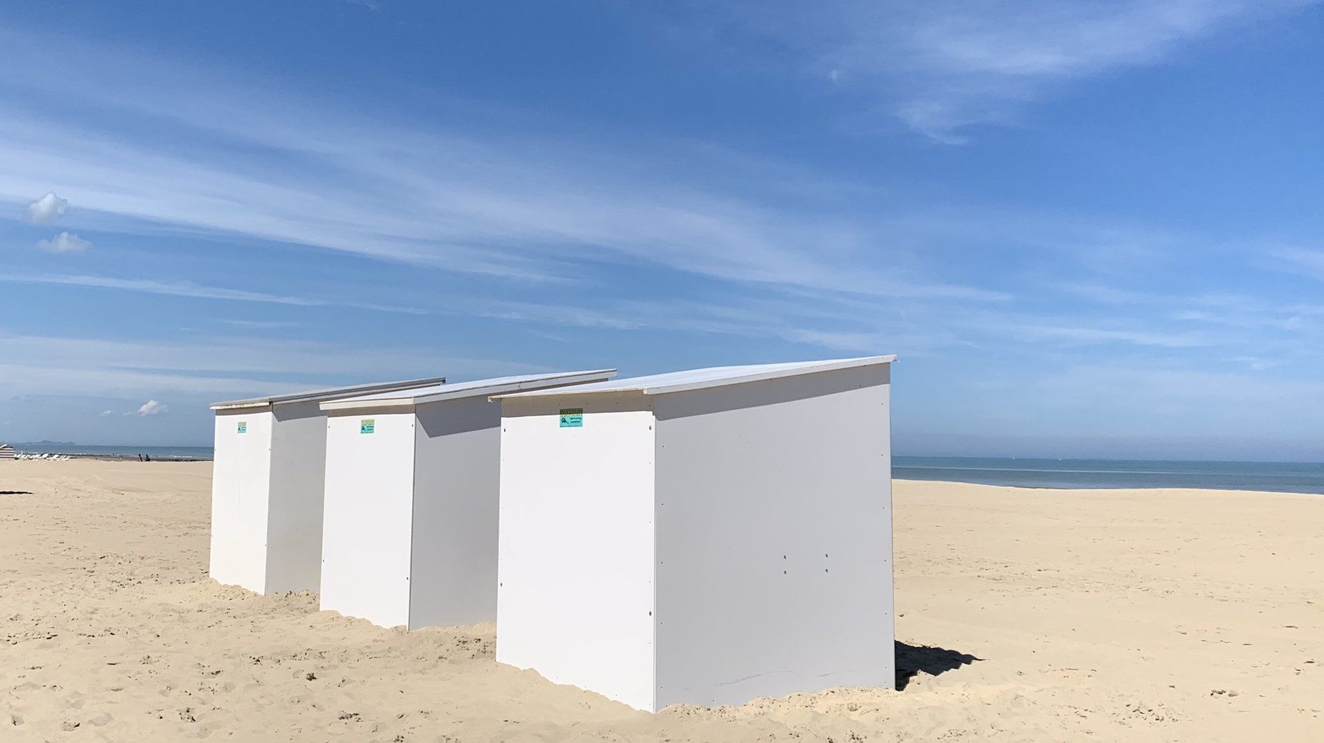 Drei weiße Strandhütten stehen aufgereiht an einem Sandstrand.