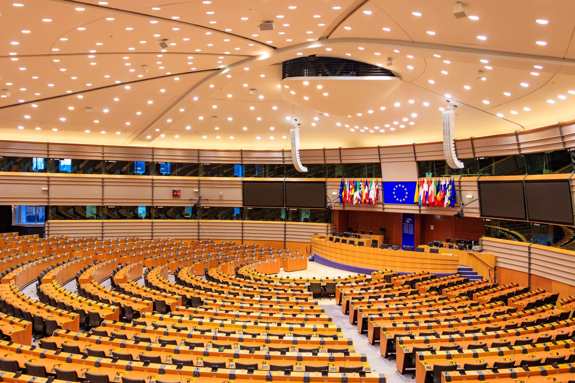 European Parliament chamber: rows of wooden seats facing a stage with flags, under a domed ceiling.