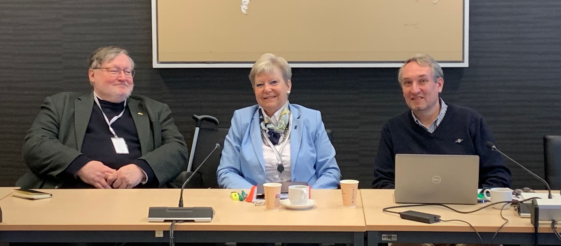 Drei Personen sitzen mit Mikrofonen und Laptops an einem Tisch.