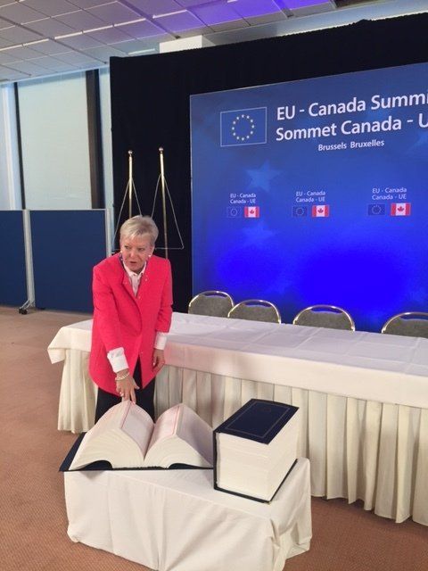 Eine Frau in einer roten Jacke steht vor einer blauen Wand mit der Aufschrift „EU Kanada Sommet Kanada“