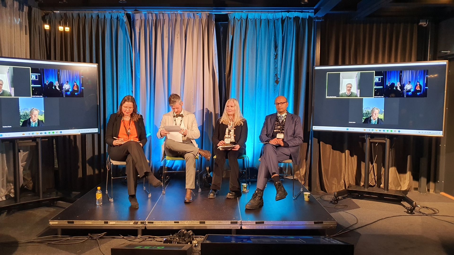 Eine Gruppe von Menschen sitzt auf einer Bühne vor zwei Fernsehern.