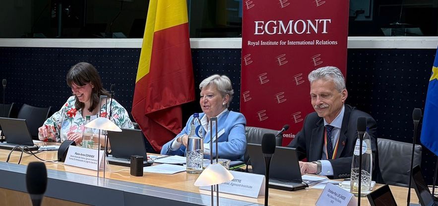 Drei Personen sitzen mit Laptops und Dokumenten an einem Tisch unter einer belgischen Flagge und einem roten Schild.