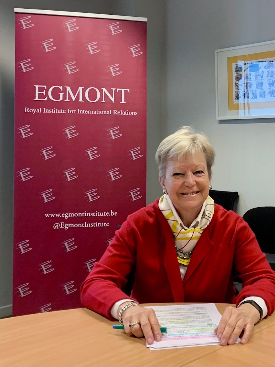 Eine Frau in einem roten Blazer sitzt lächelnd an einem Schreibtisch vor einem EGOMNT-Banner und hält einen Stift über ihre Blätter.