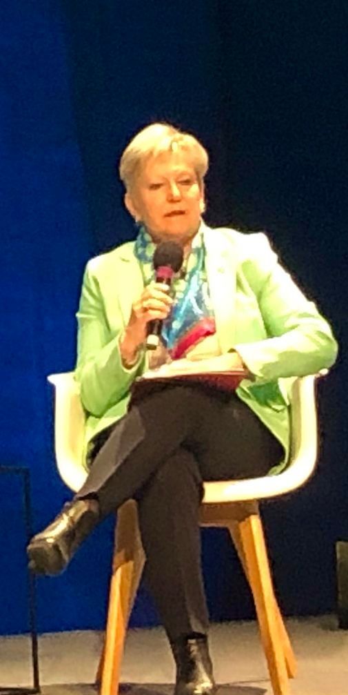 Eine Frau in einer grünen Jacke sitzt auf einem Stuhl und hält ein Mikrofon.