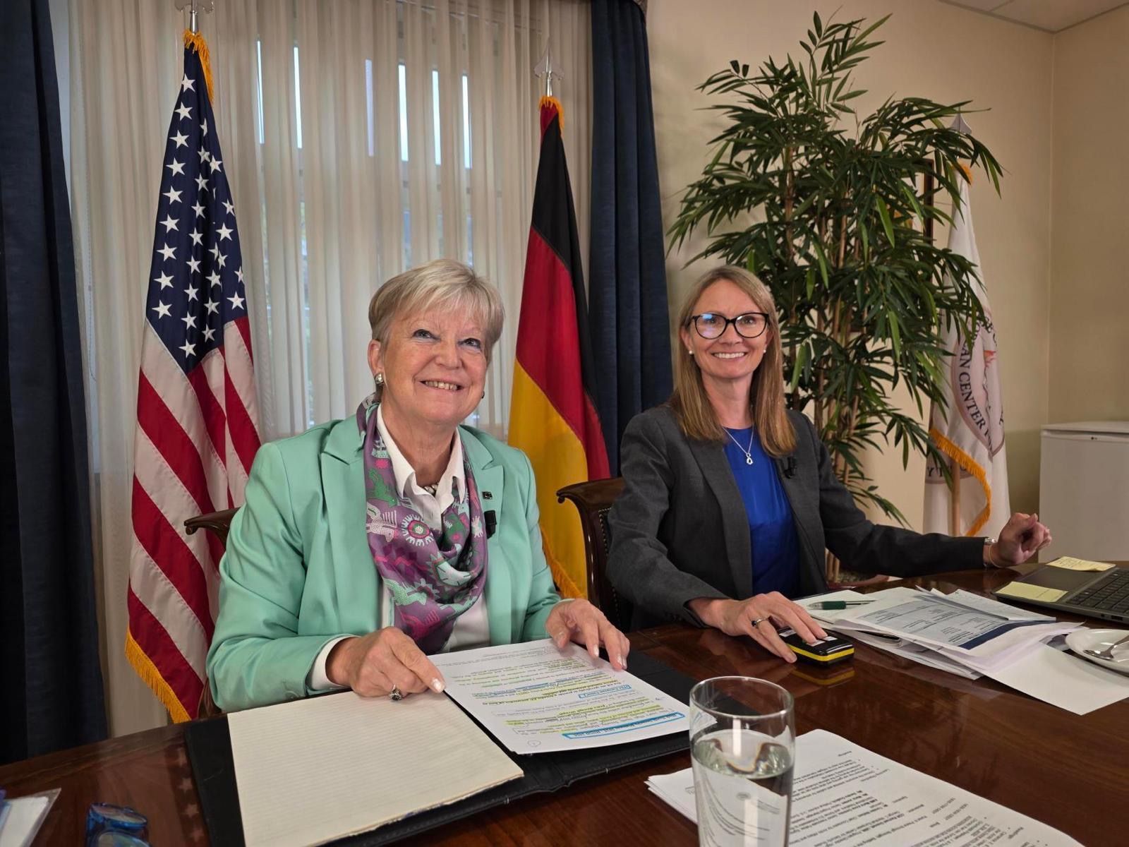 Zwei Frauen sitzen lächelnd an einem Schreibtisch. Hinter ihnen die US-amerikanische und die deutsche Flagge.
