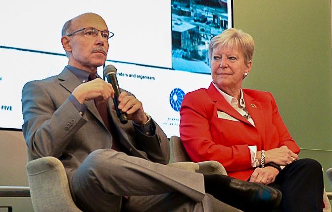 Ein Mann im Anzug spricht in ein Mikrofon und sitzt neben einer Frau in einer roten Jacke.