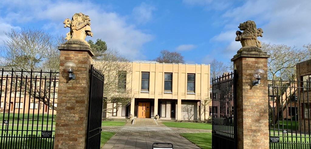 Hinter einem Tor mit einem Löwen darauf befindet sich ein großes Gebäude.