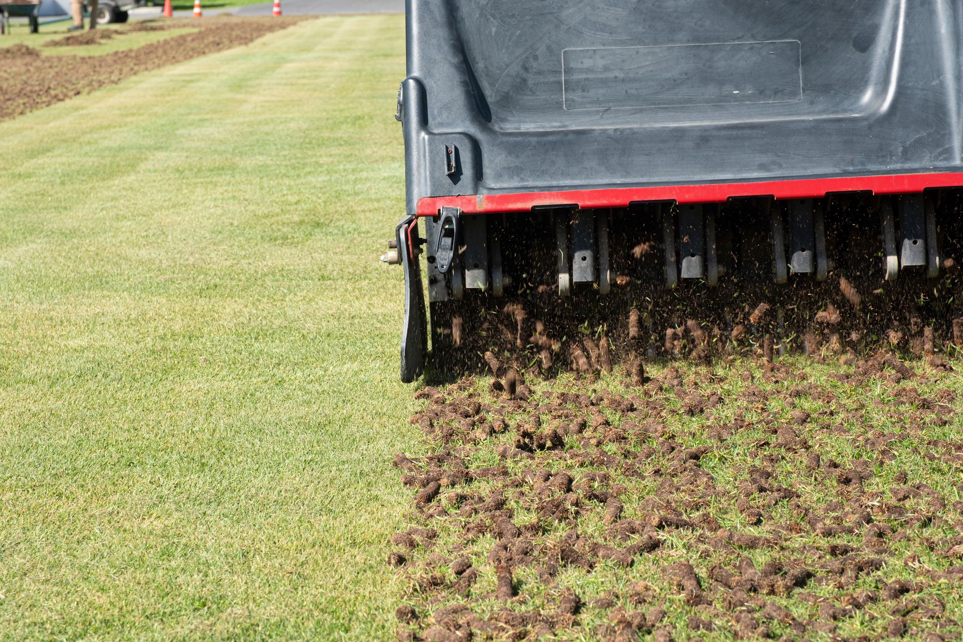 A lawn aerator machine moves across green grass, leaving behind small, cylindrical plugs of soil on the turf.