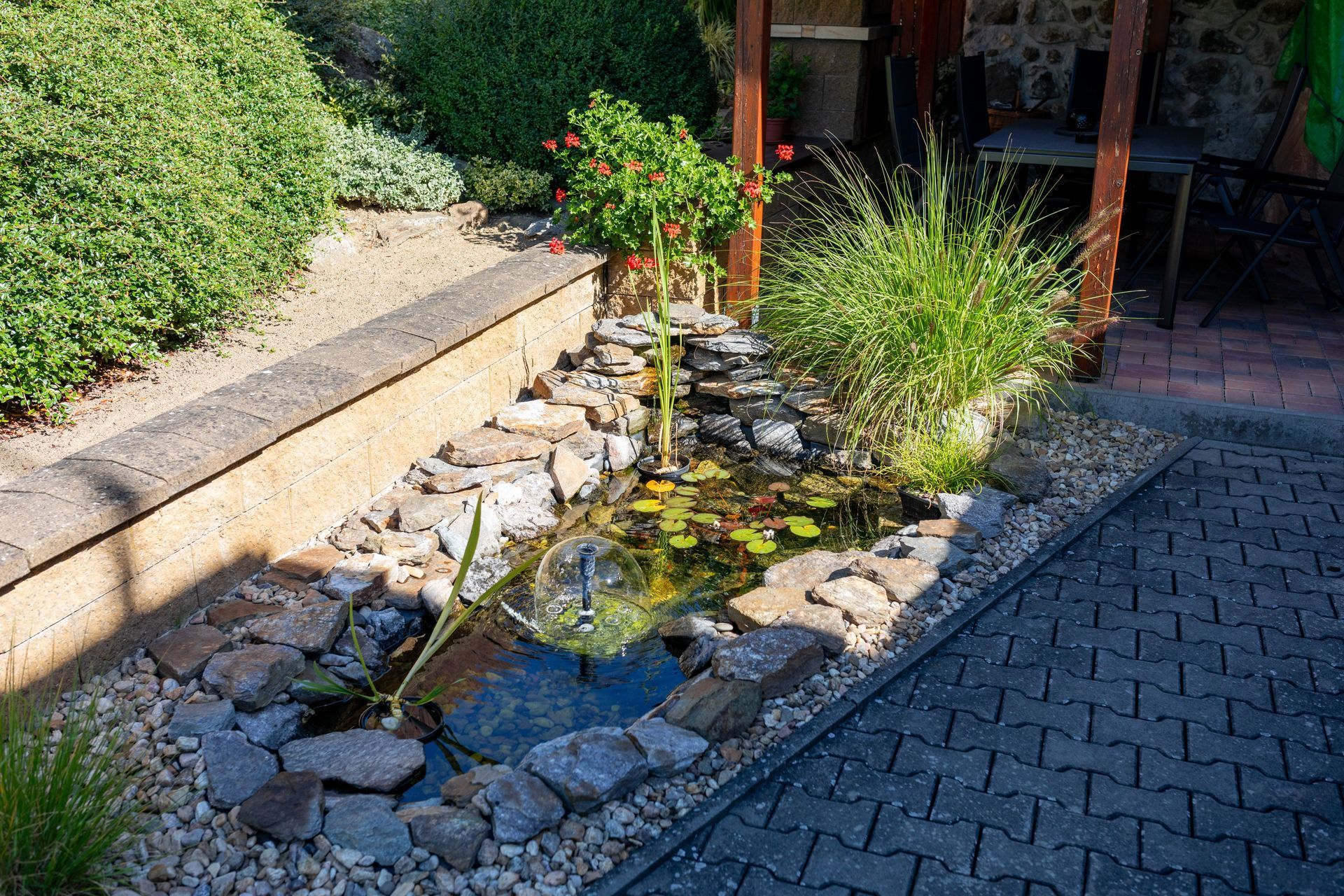 A stone-bordered backyard pond with water lilies and a small fountain, next to a patio area and garden bushes.