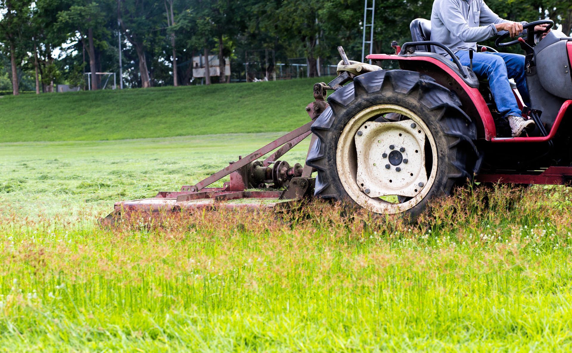 A person driving a red tractor pulls a mower through a green field, cutting the tall grass.
