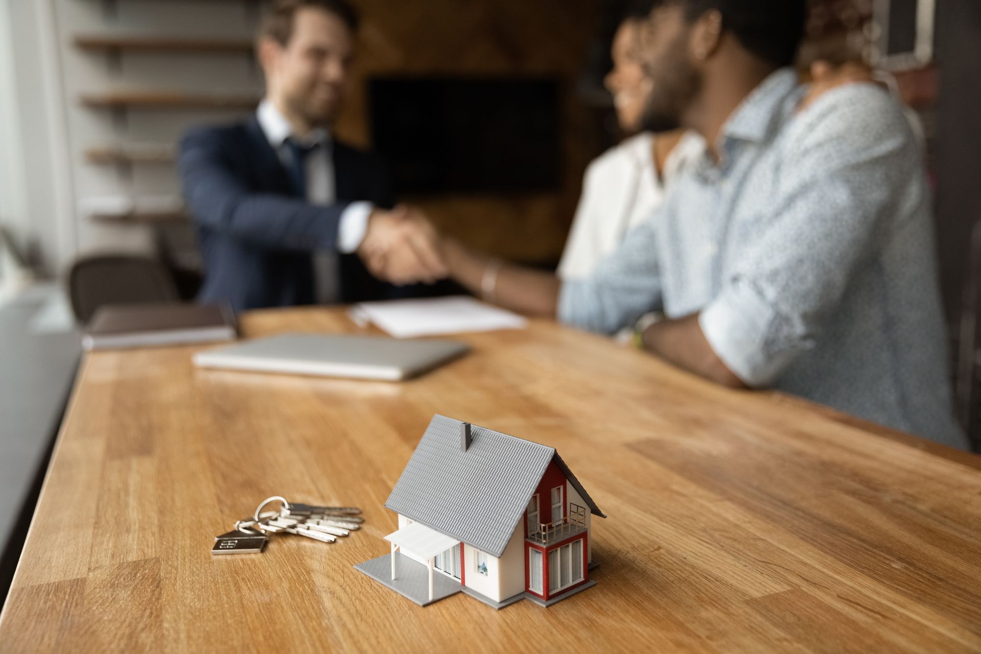 A man and woman are shaking hands over a table with a model house and keys.