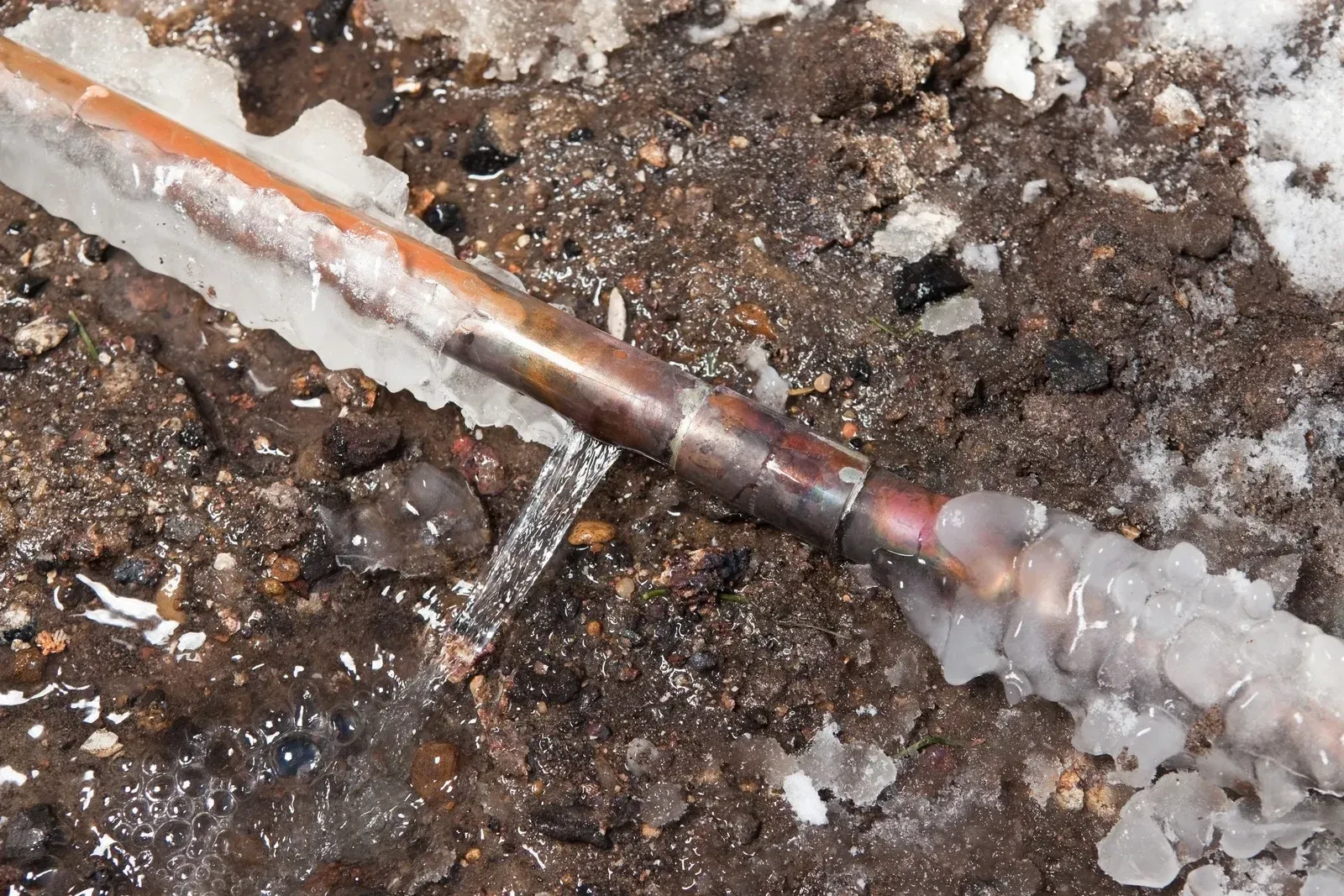 Frozen and burst copper pipe leaking water onto dark soil with snow.