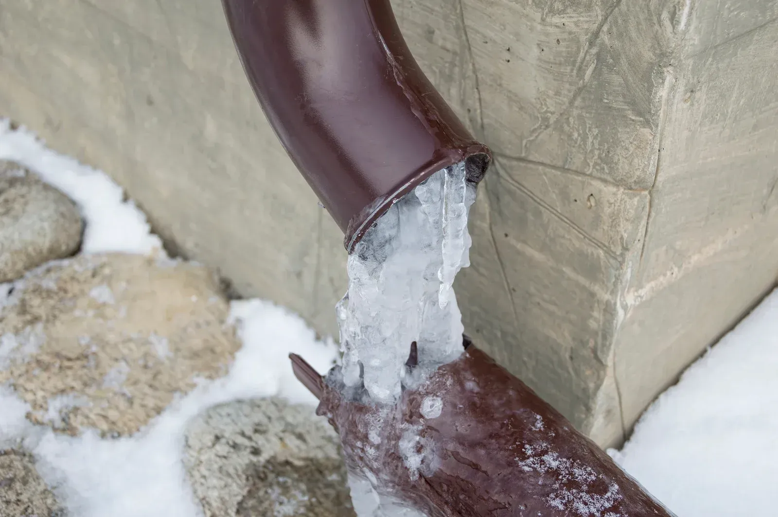 Ice buildup blocking a brown gutter spout next to a concrete wall and snow-covered rocks.
