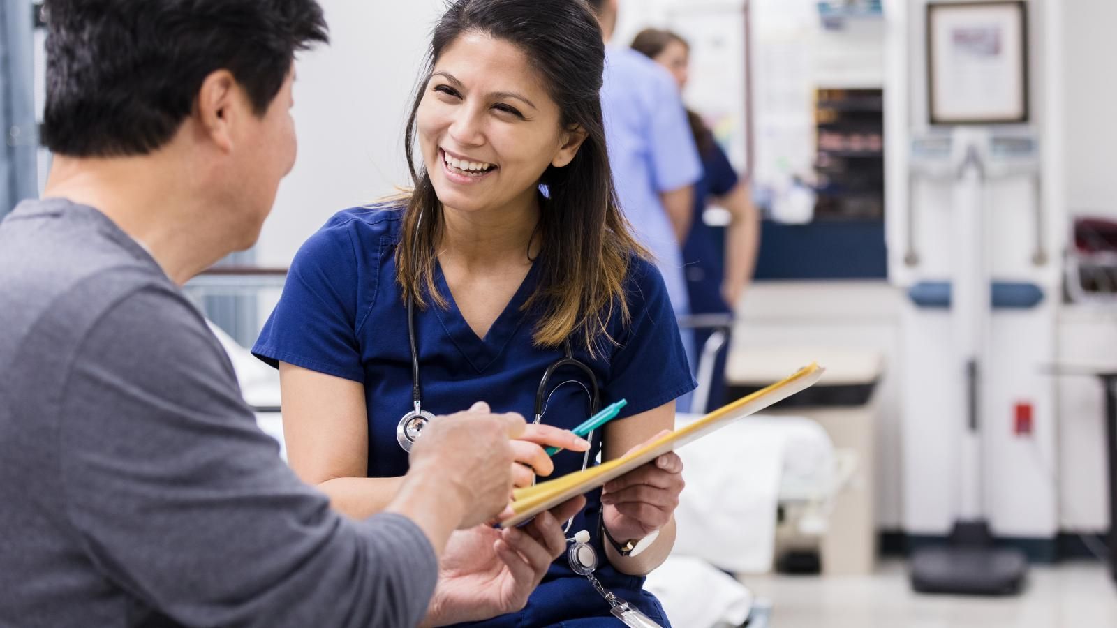 A nurse is talking to a patient in a hospital room while holding a clipboard.