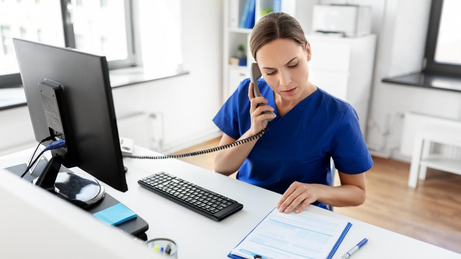 A nurse is sitting at a desk talking on a phone in front of a computer.