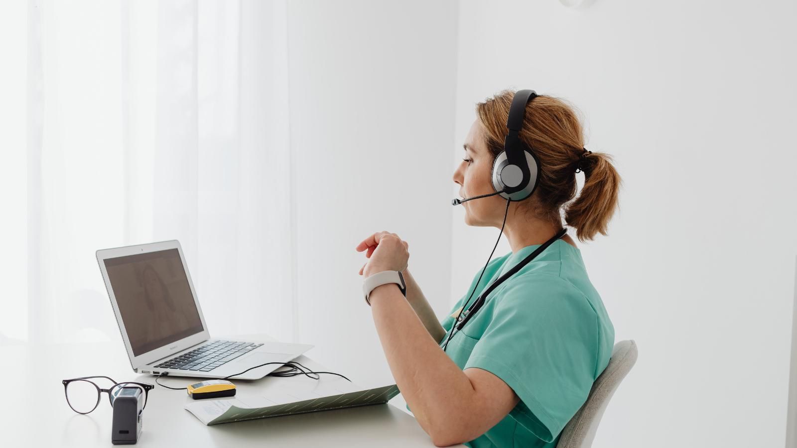 Medical professional in scrubs with headset on video call via laptop at desk.