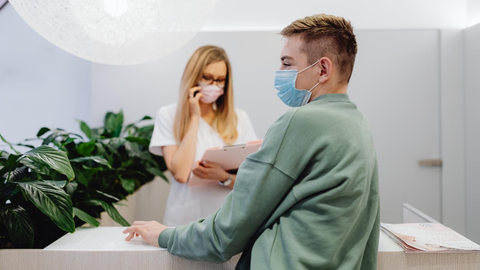 Person at a reception desk, wearing a face mask. Another person in background on the phone. Green plants.