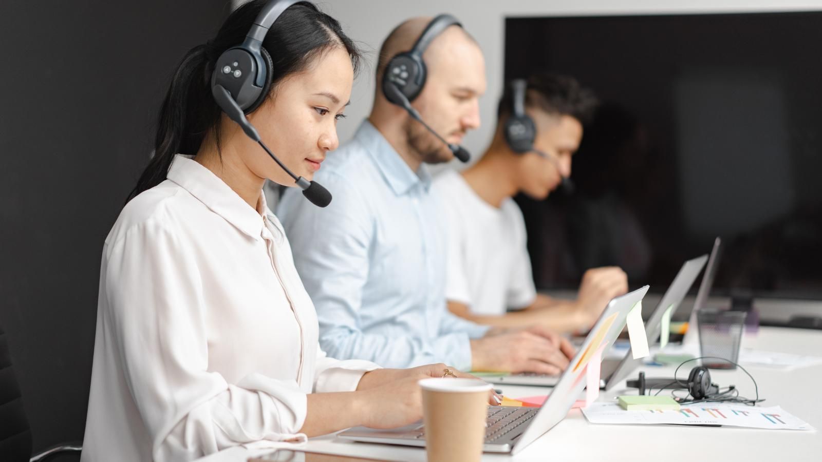 A group of virtual receptionist wearing headsets are sitting at a table with laptops.