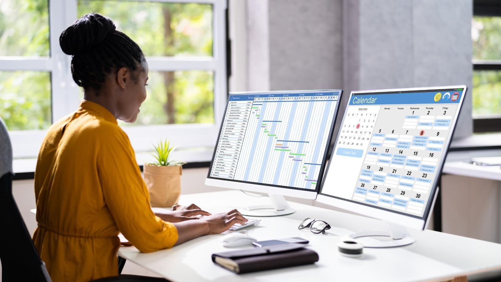 A woman is sitting at a desk in front of two computer monitors, scheduling patient appointments.