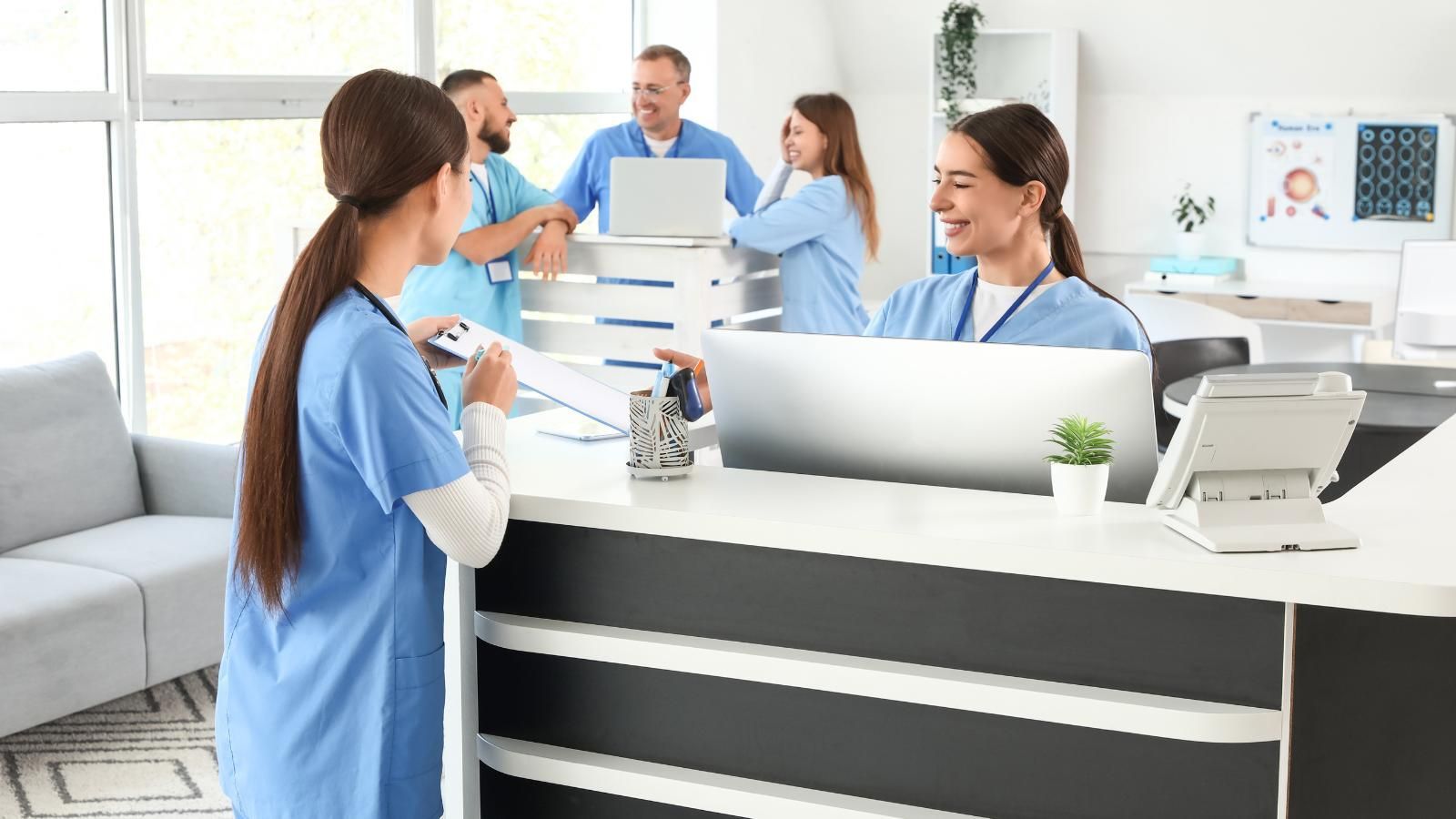 strategies to improve patient intake efficiency Medical staff at a reception desk, one taking notes, others behind the counter talking near laptops, bright office.