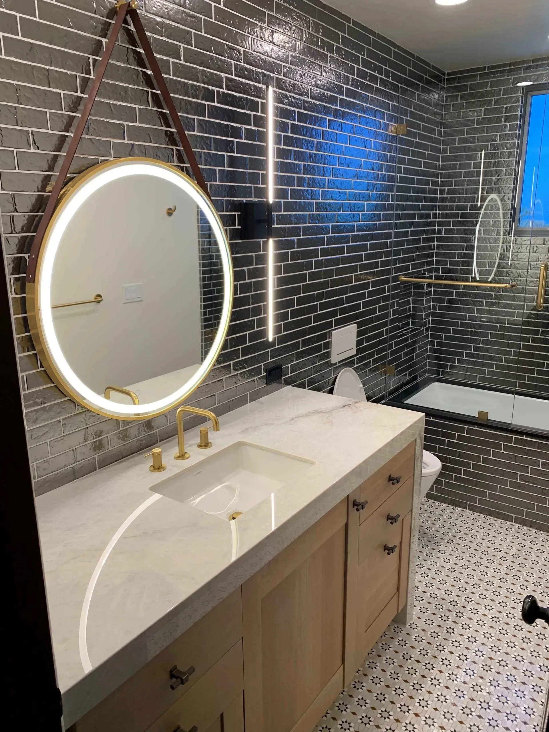 Modern bathroom with a round mirror, gold fixtures, marble countertop, and black brick-like wall tiles.