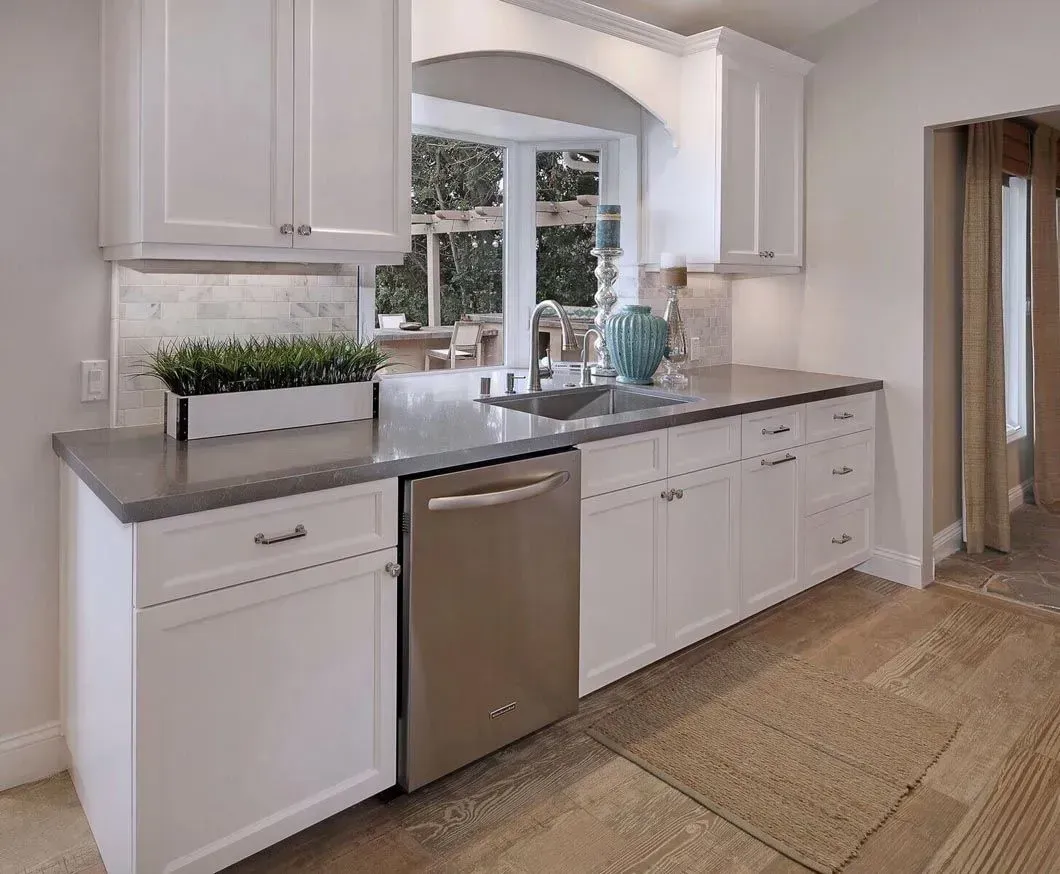 White kitchen with stainless steel dishwasher and grey countertop, overlooking an outdoor patio.
