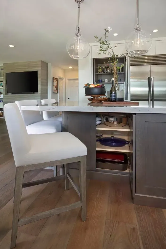 Kitchen island with white chairs, gray cabinets, and built-in shelving, with overhead lighting.