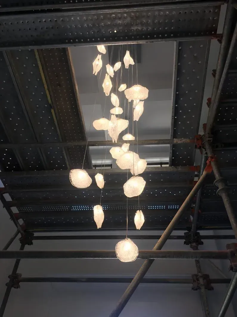 Chandelier with glowing, organic-shaped lights hanging from the ceiling, viewed from below. Scaffolding surrounds it.