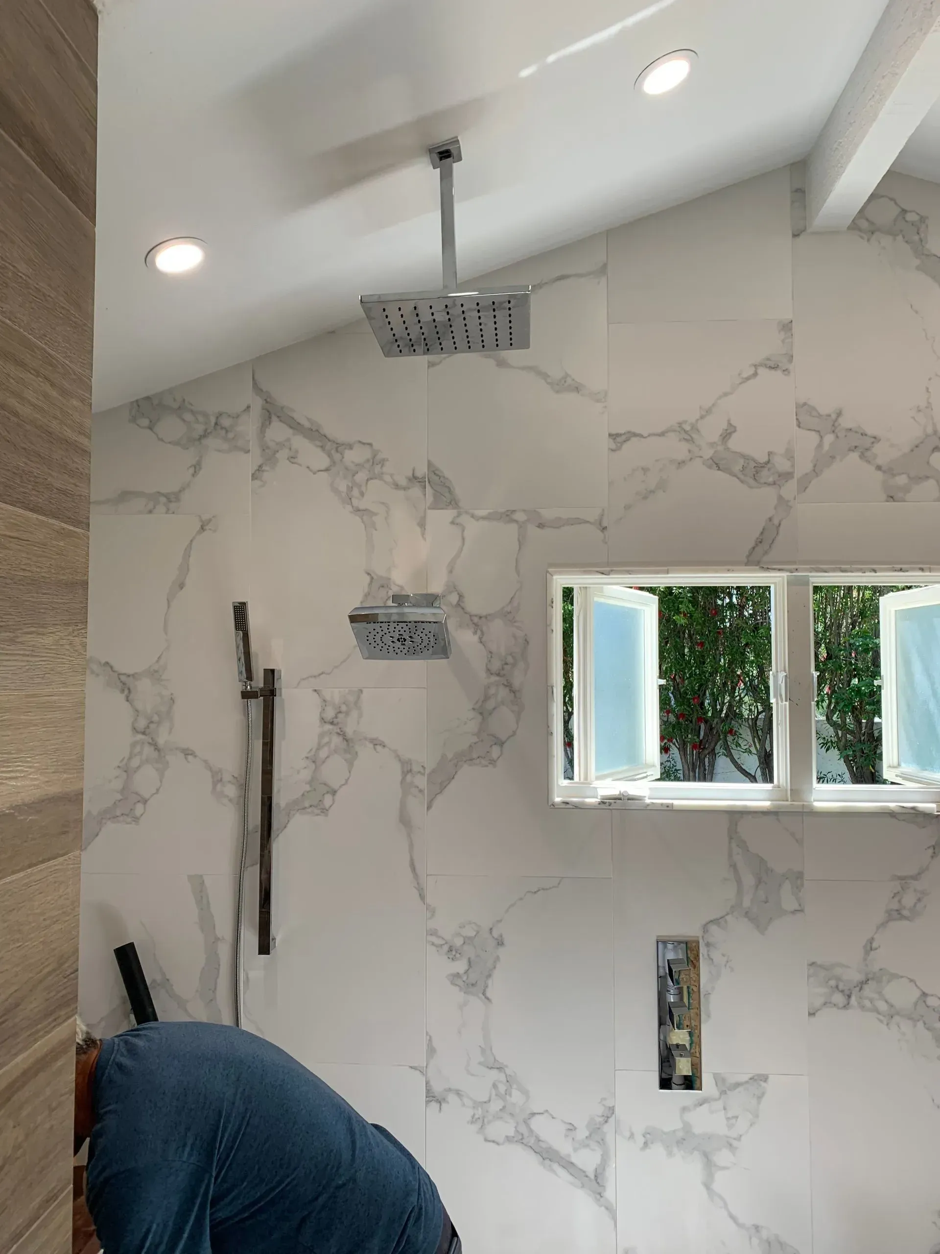 Bathroom shower with marble-like wall tile, window, and a person working.