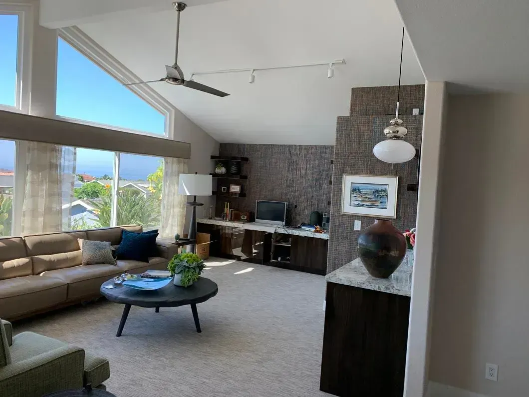 Living room with high ceilings, large windows, and a sectional sofa; desk and shelves are visible.